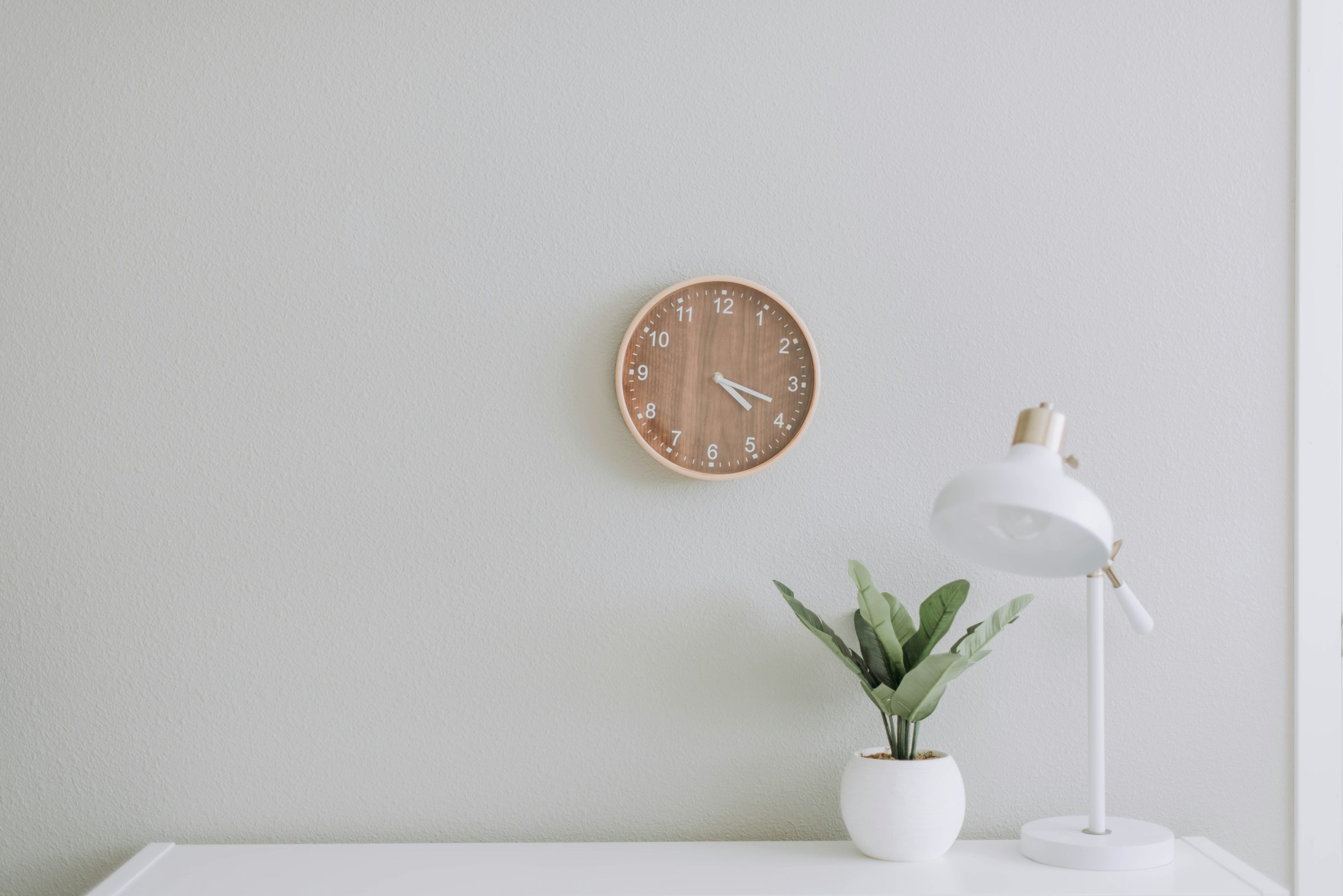 White desk lamp and plant in front of a white wall with a clock